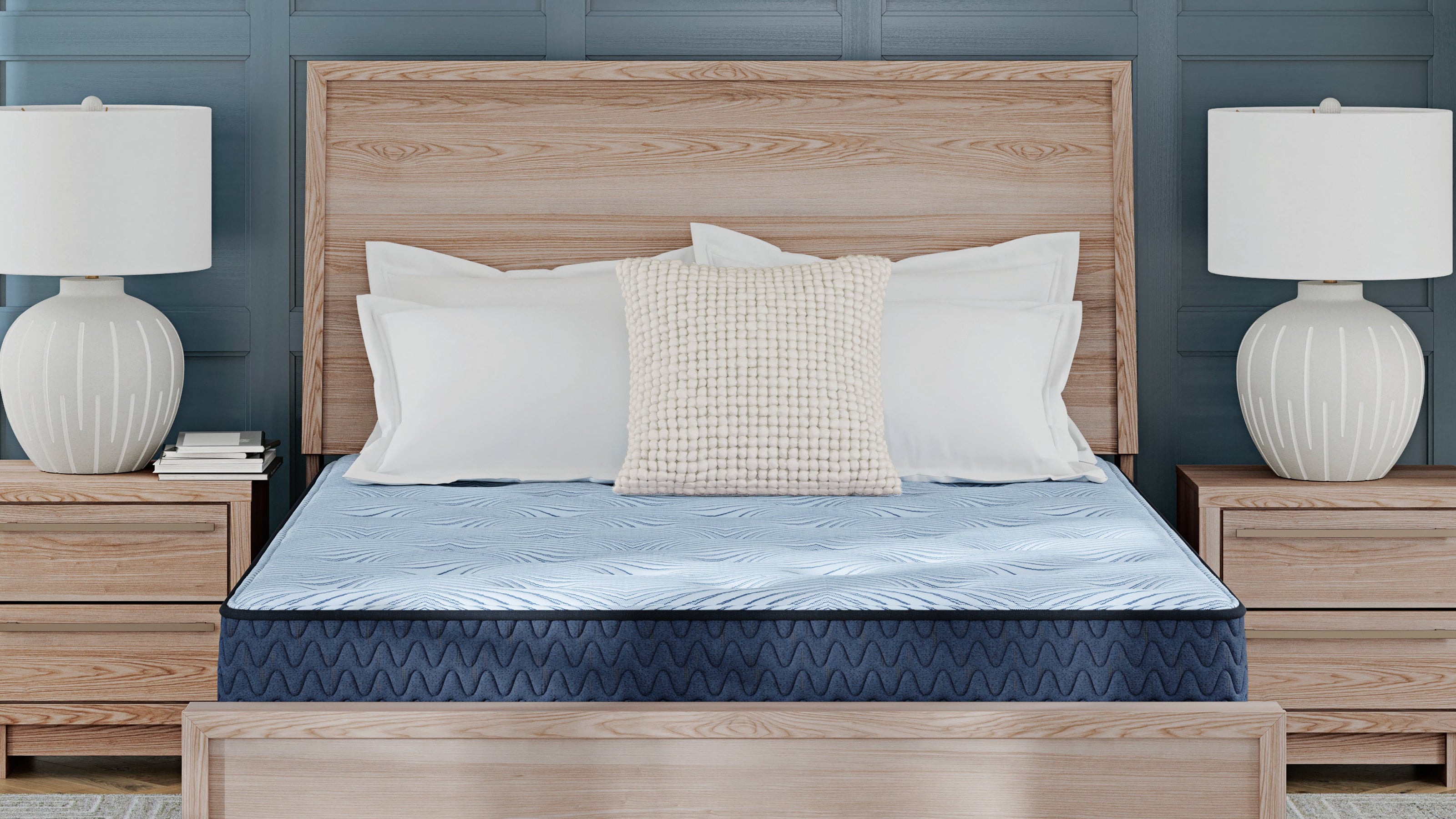 A light brown wooden bed frame with a matching headboard and footboard, styled with a blue patterned mattress, white pillows, and flanked by matching nightstands with white table lamps against a teal paneled wall.