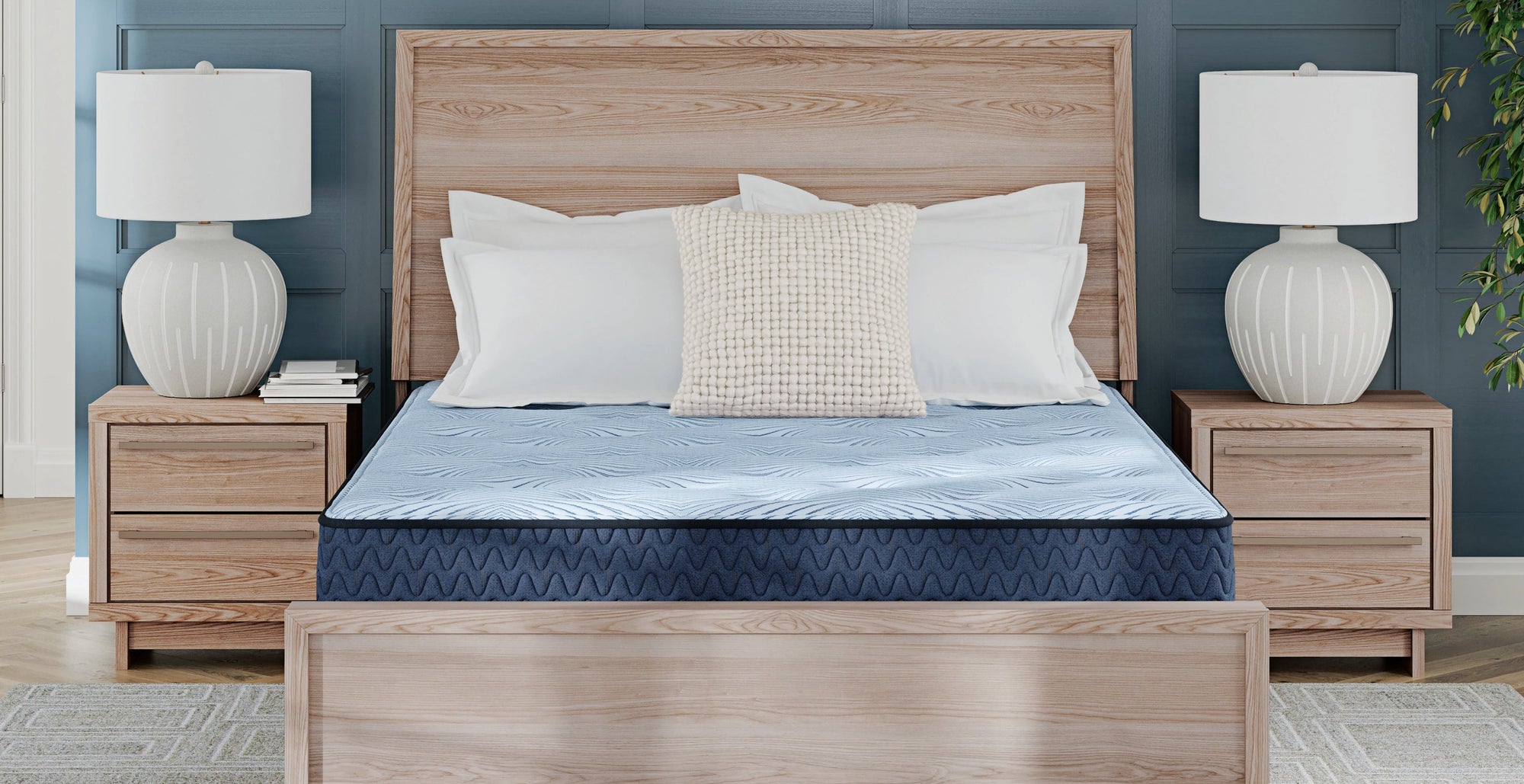 A light oak wood bed frame with a matching headboard and footboard, styled with a blue patterned mattress, white pillows, and a textured cream pillow, flanked by matching nightstands and white table lamps against a blue-gray wall.