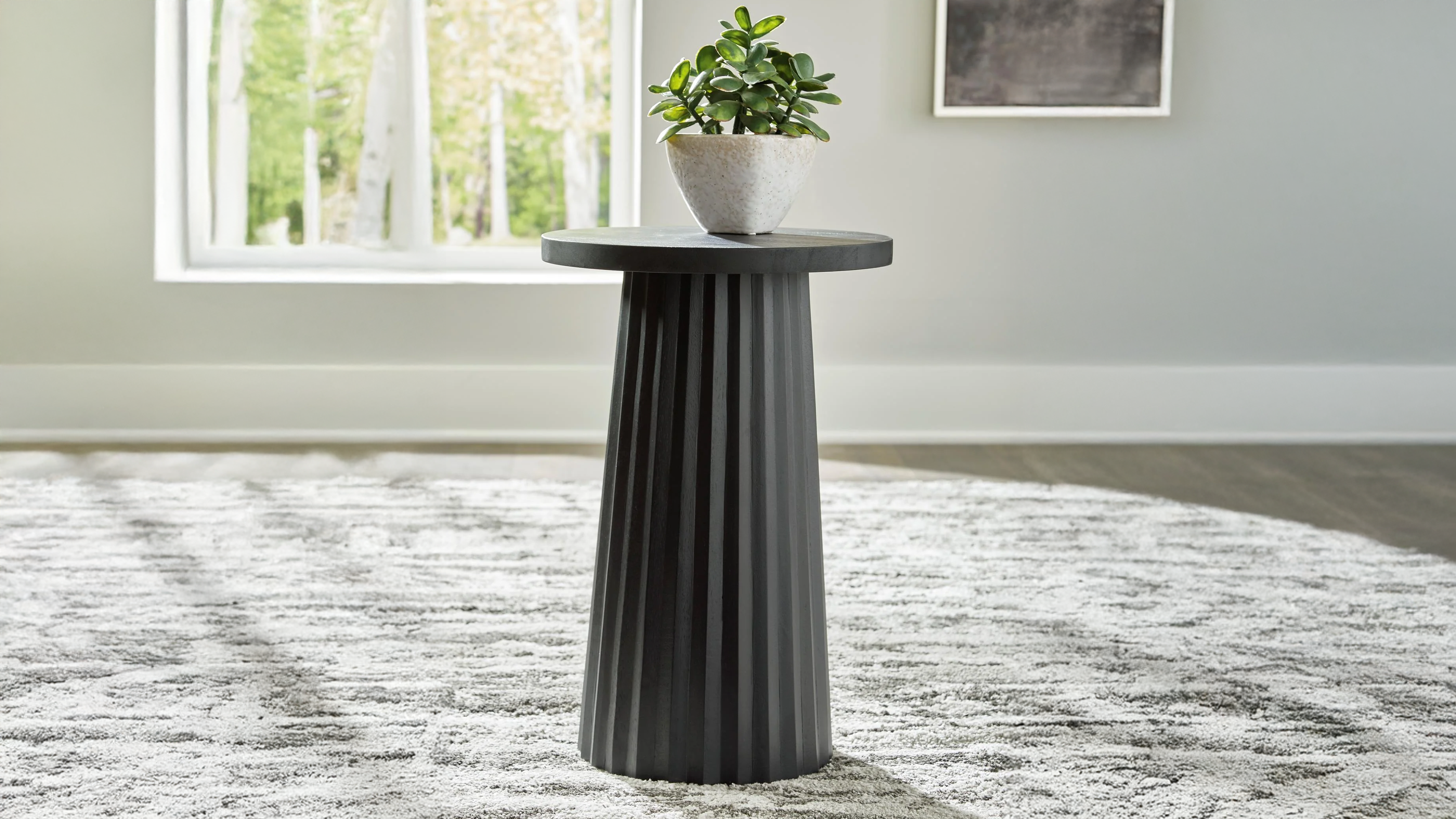 Black cylindrical side table with a plant on a gray rug in a room with a window and wall art.