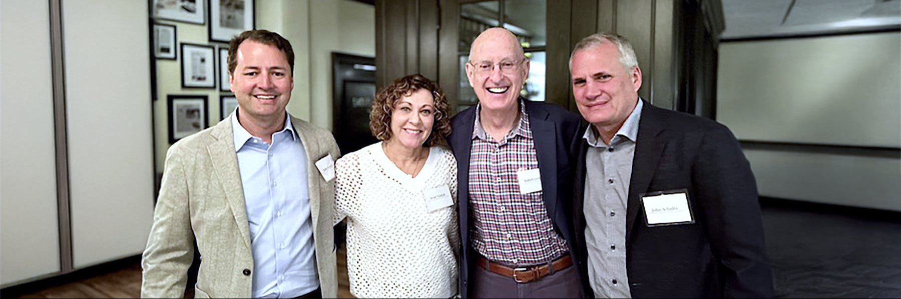 Four people standing together in an indoor hallway or office setting, wearing business casual attire and name tags.