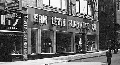Black and white photograph of a historic storefront for Sam Levin Furniture Co., with large window display windows, a street sidewalk, and a pedestrian visible in the lower right.