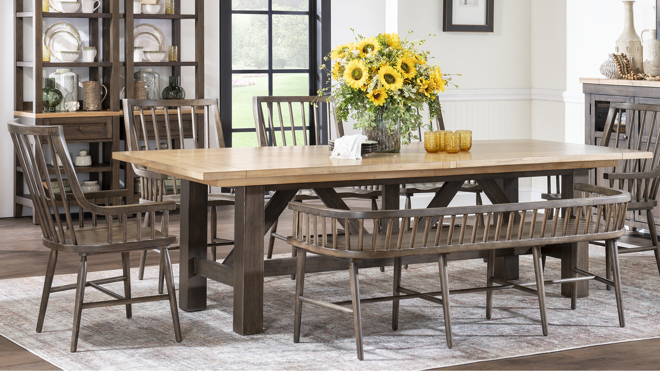 A rustic dining room set featuring a rectangular light oak wood table with dark brown trestle-style legs, surrounded by matching wooden chairs and a bench on a patterned rug. A vase of yellow sunflowers sits on the table, and the background shows a styled home interior with shelving and a sideboard.