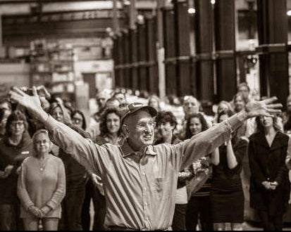 A black and white photograph of a man with arms outstretched, smiling and celebrating in a crowd of people inside a large warehouse.
