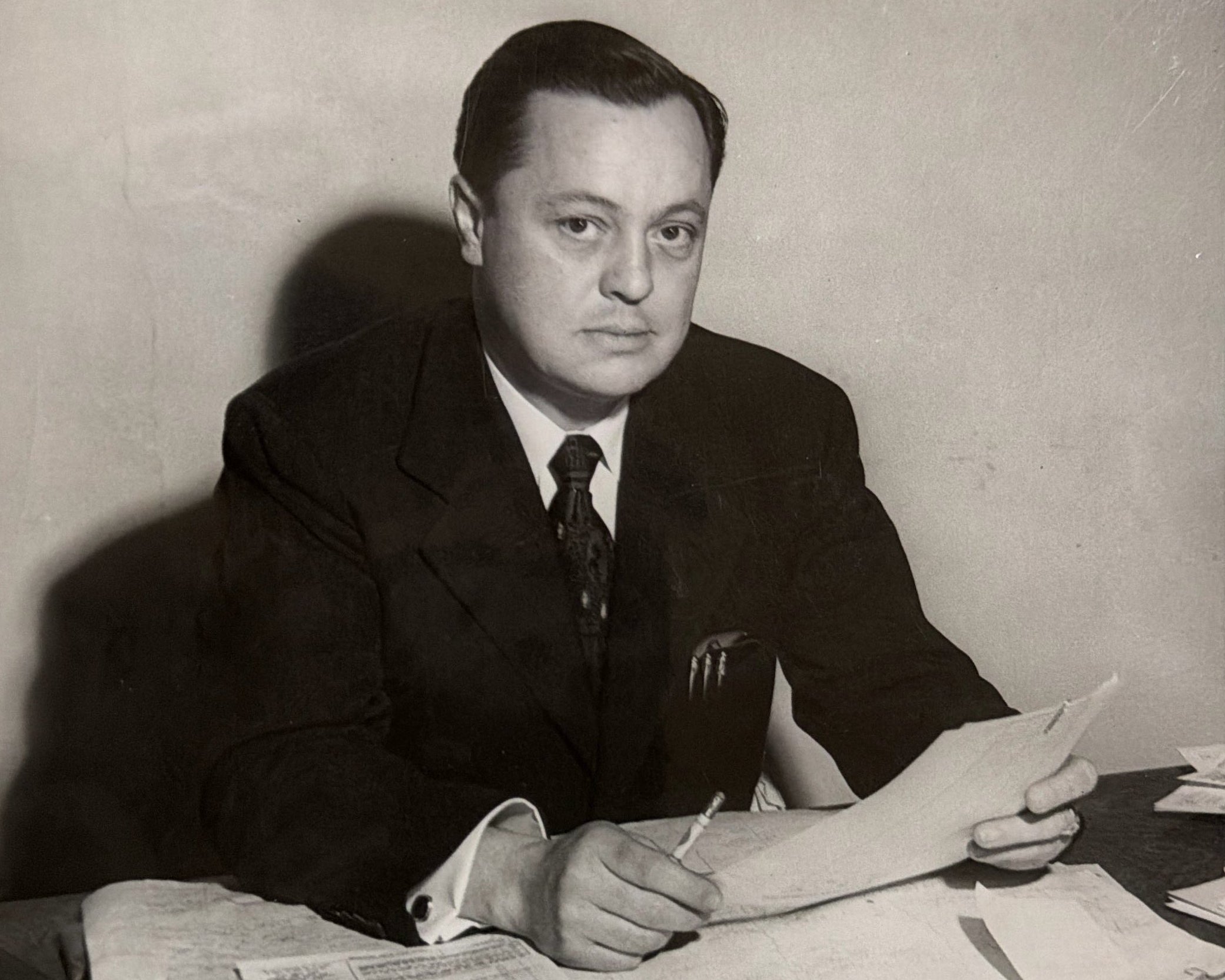 A vintage black and white photograph of a man in a dark suit and tie sitting at a desk surrounded by papers.