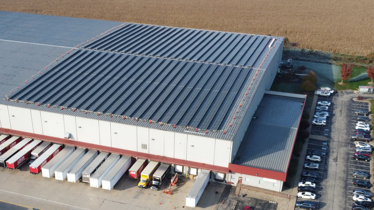 An aerial view of a large industrial warehouse with a dark metal roof covered in solar panels, loading dock bays with semi-trucks, and a parking lot on the right.