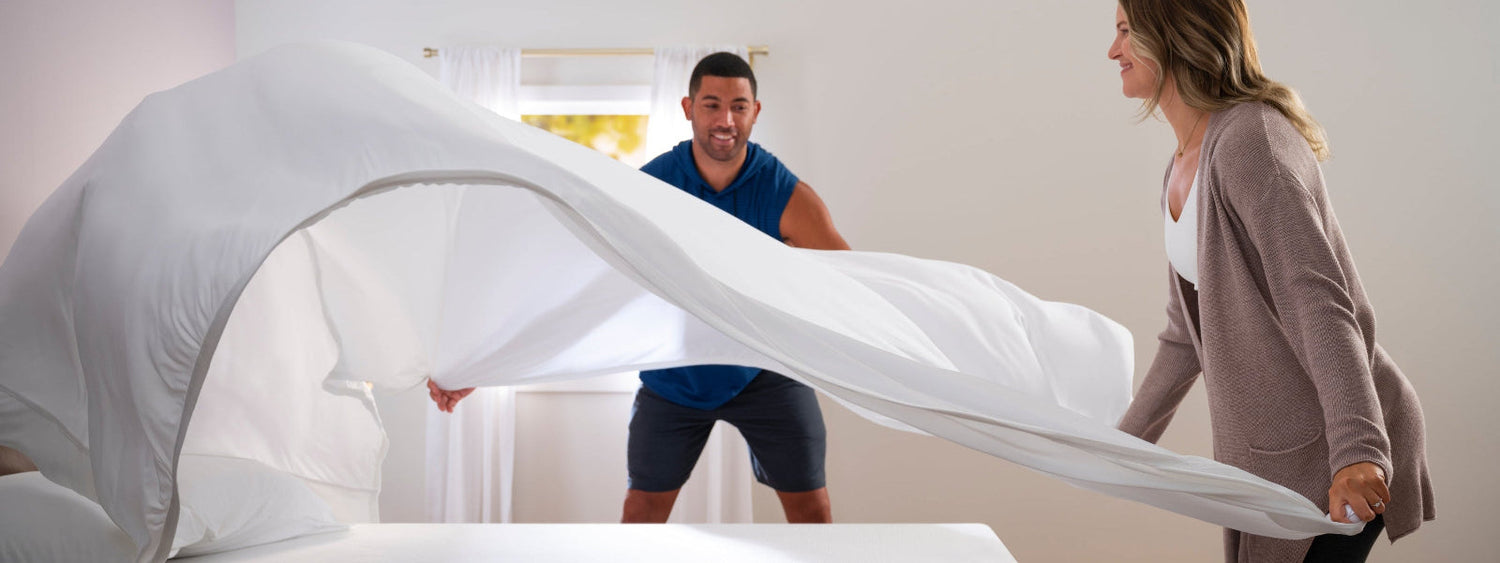 Two people setting up a bed with white bedding in a bright room.