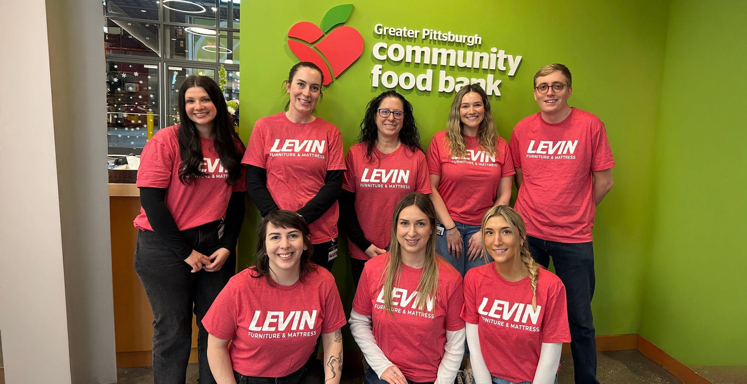 A group of eight people wearing matching red t-shirts with 'LEVIN' printed on the front, standing together in front of a green wall with the Greater Pittsburgh Community Food Bank logo.
