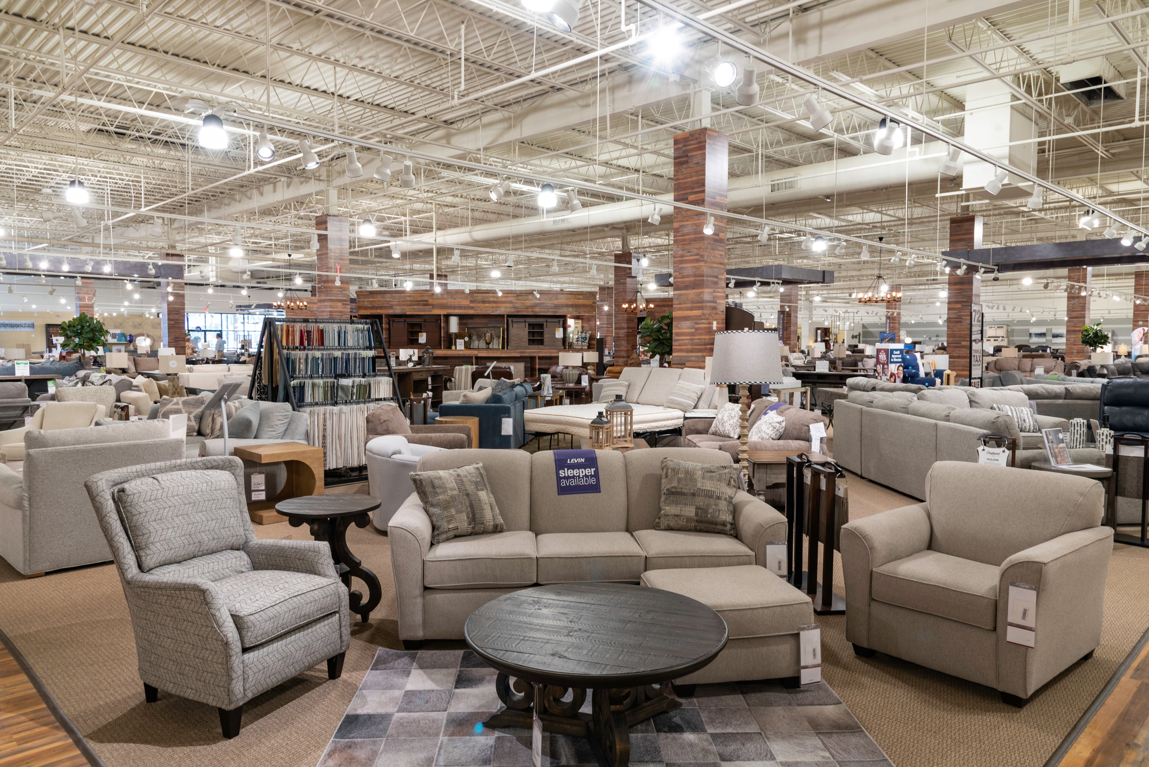 A wide showroom interior with multiple upholstered seating pieces, tables, lamps, and shelving. In the foreground, a light beige sofa is centered with two matching armchairs, a round dark wood coffee table, and patterned area rugs on a large retail floor.