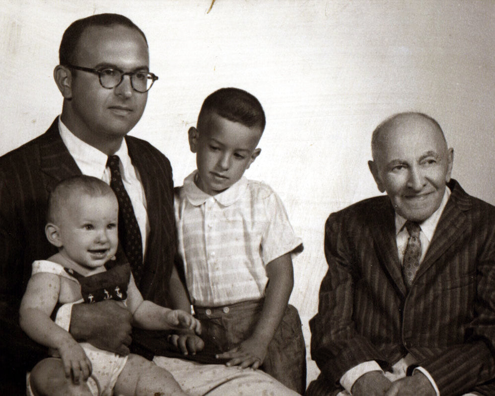 A vintage black and white family portrait showing four people: an older man on the right, a younger man on the left holding a baby, a young boy in the center, and an older man seated next to them against a plain light background.